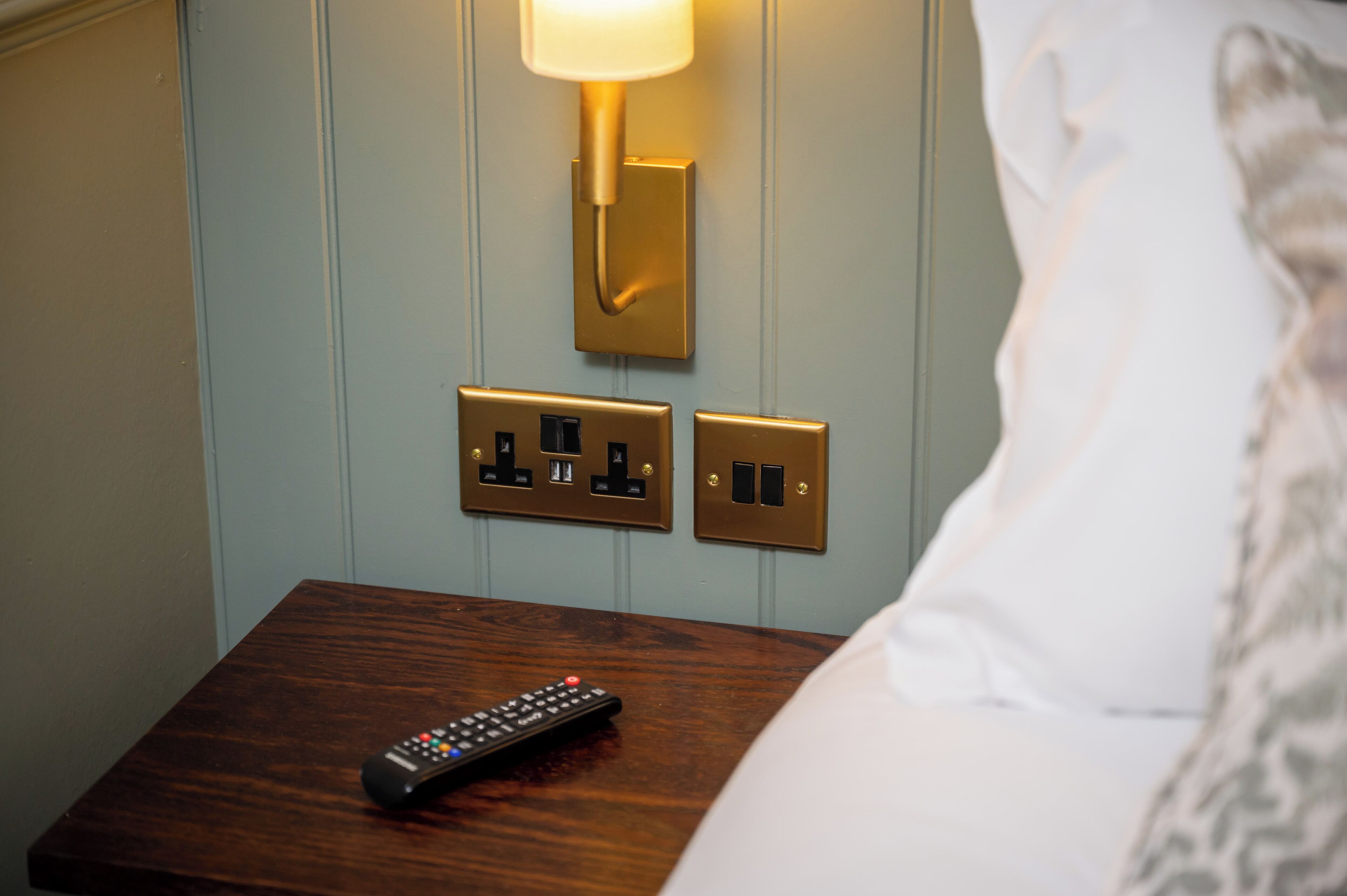 A wooden bedside table with a remote control, a wall-mounted light fixture, and electrical outlets.