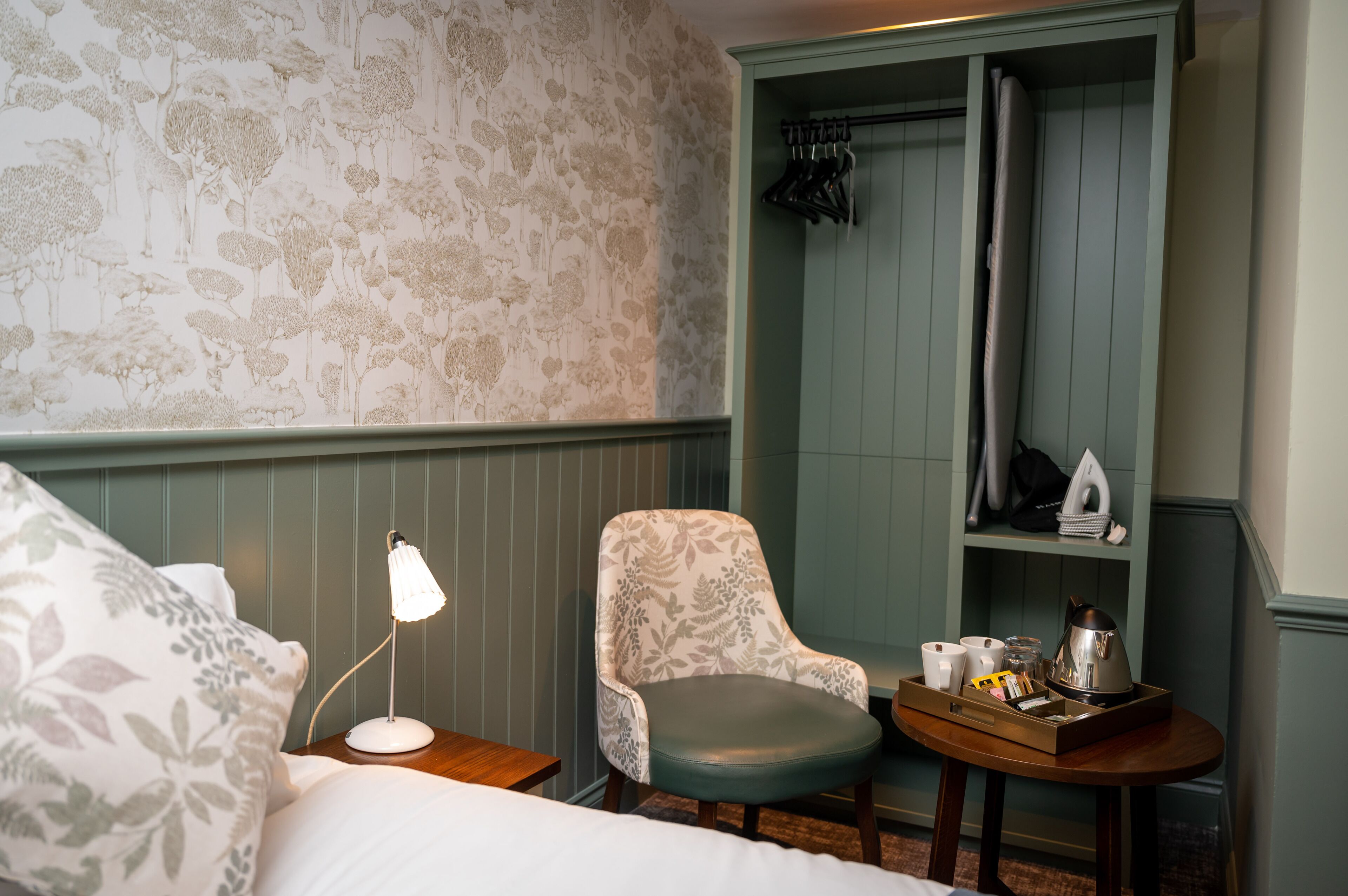 A neatly arranged hotel room with a bed, a chair, a small table with a tray, and a wardrobe with hanging clothes.