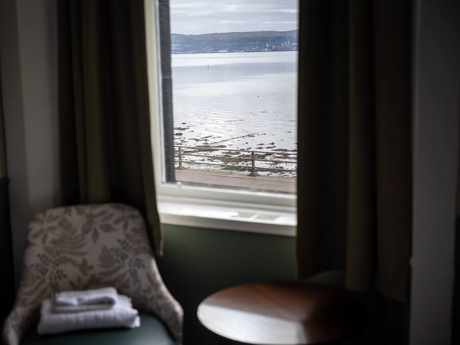 Quarto com vista para um corpo d'água através de uma janela, uma cadeira com toalha dobrada e uma pequena mesa.