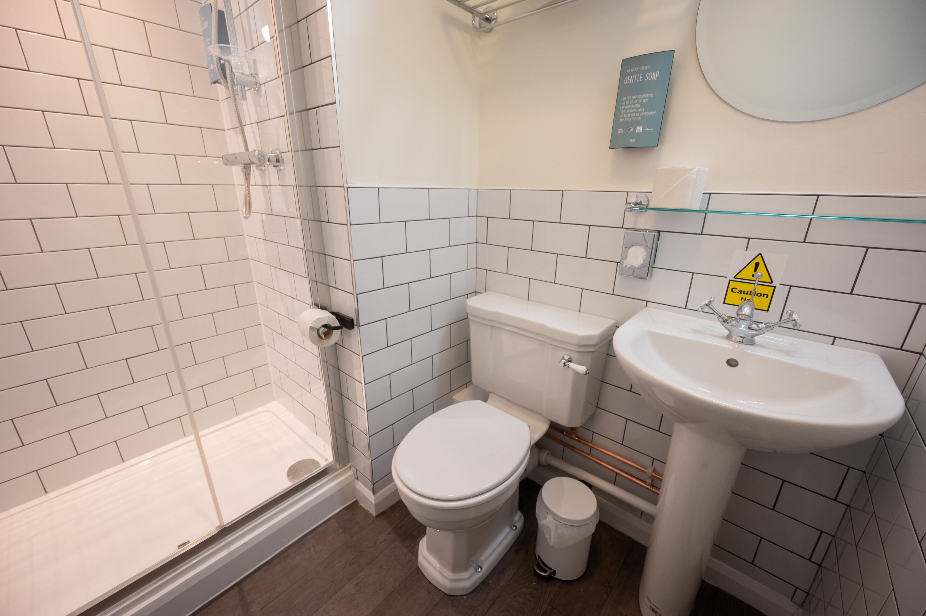 A bathroom with a glass shower enclosure, a white toilet, and a sink with a caution sign.