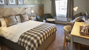 Een hotelkamer met een groot bed, een bureau met een lamp, een stoel, een klein rond tafeltje en een raam met gordijnen.