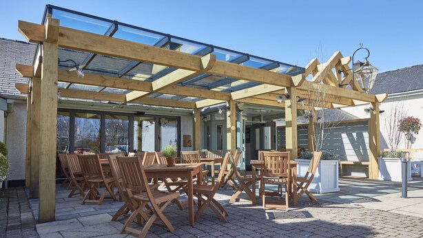 Een terras buiten met houten tafels en stoelen, een glazen overkapping en potplanten.