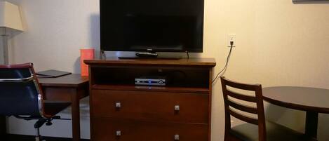 Habitación de hotel con televisor de pantalla plana sobre un soporte de madera, un escritorio con silla, un mueble pequeño y una mesa redonda.