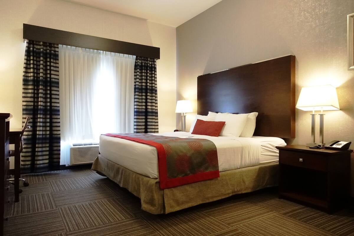 Une chambre d’hôtel avec un grand lit, un bureau et une chaise. Il y a une fenêtre avec des rideaux et une lampe sur la table de chevet.