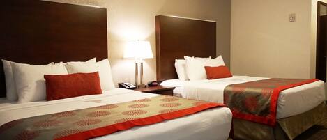 Habitación de hotel con dos camas, cada una con ropa blanca y detalles rojos, una mesita de noche con lámpara y teléfono.