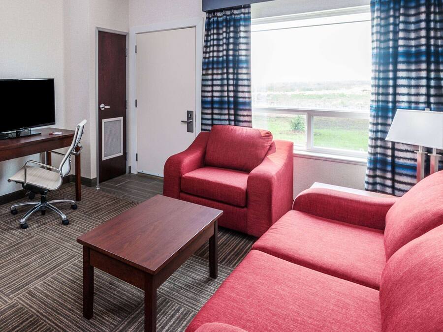 Habitación de hotel con televisión, escritorio, sillón rojo, sofá y ventana con cortinas.