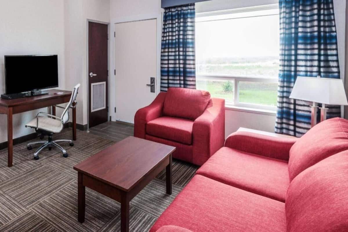 Une chambre d’hôtel équipée d’une télévision, d’un bureau, d’un fauteuil rouge, d’un canapé et d’une fenêtre avec des rideaux.