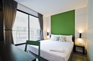 Habitación de hotel con un ventanal grande, una pared de acento verde, una cama bien hecha con ropa blanca y una silla junto a una mesa de madera.