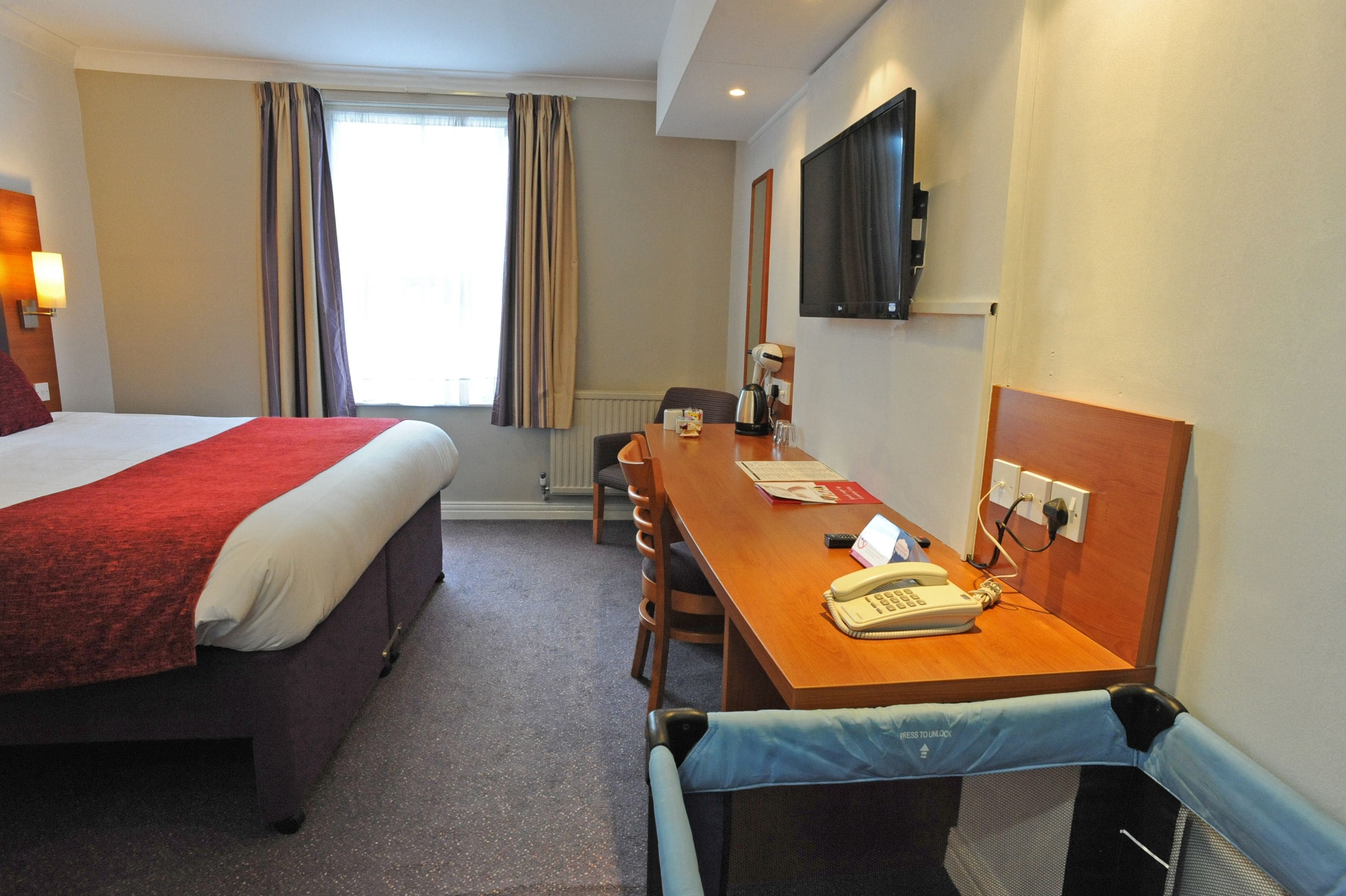 A hotel room with a large bed, a wooden desk with a telephone, a chair, and a television mounted on the wall.