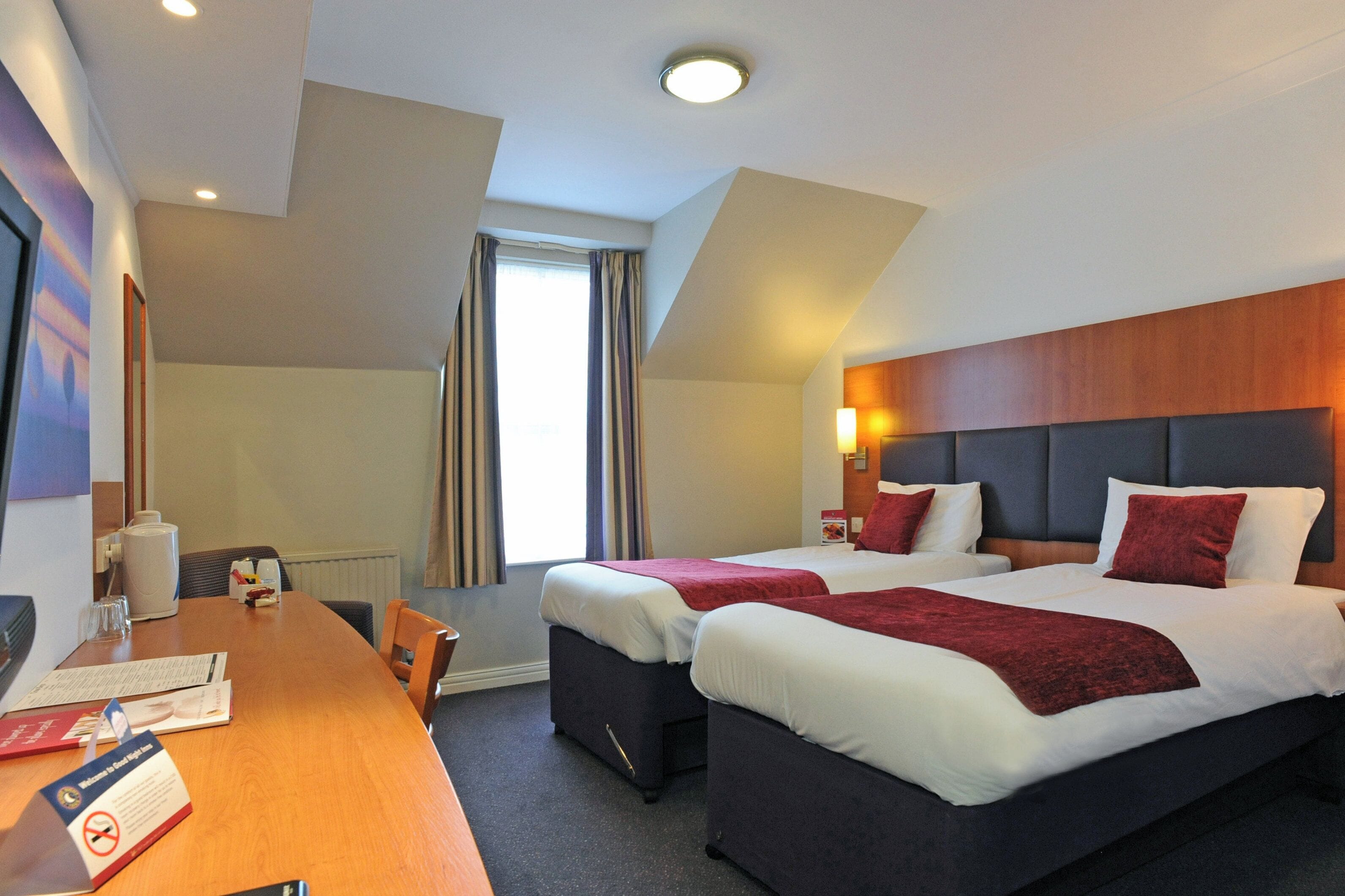 A hotel room with two beds, a wooden desk, and a view of the outdoors.