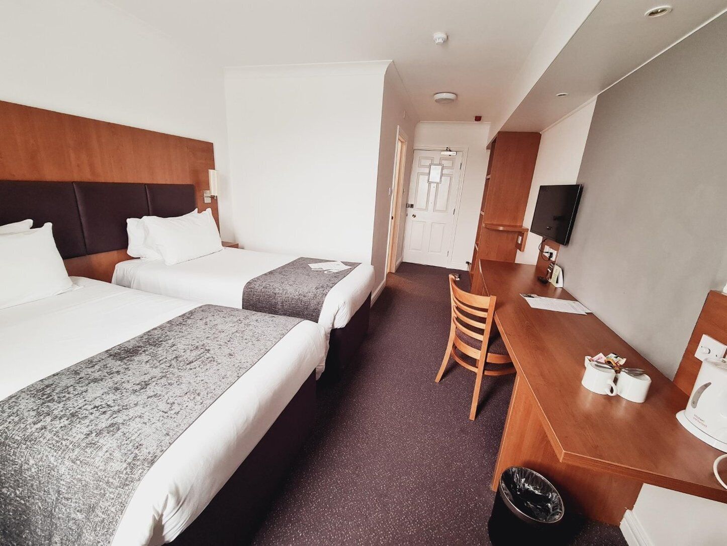 A hotel room with two beds, a desk, a chair, a TV, and a door.
