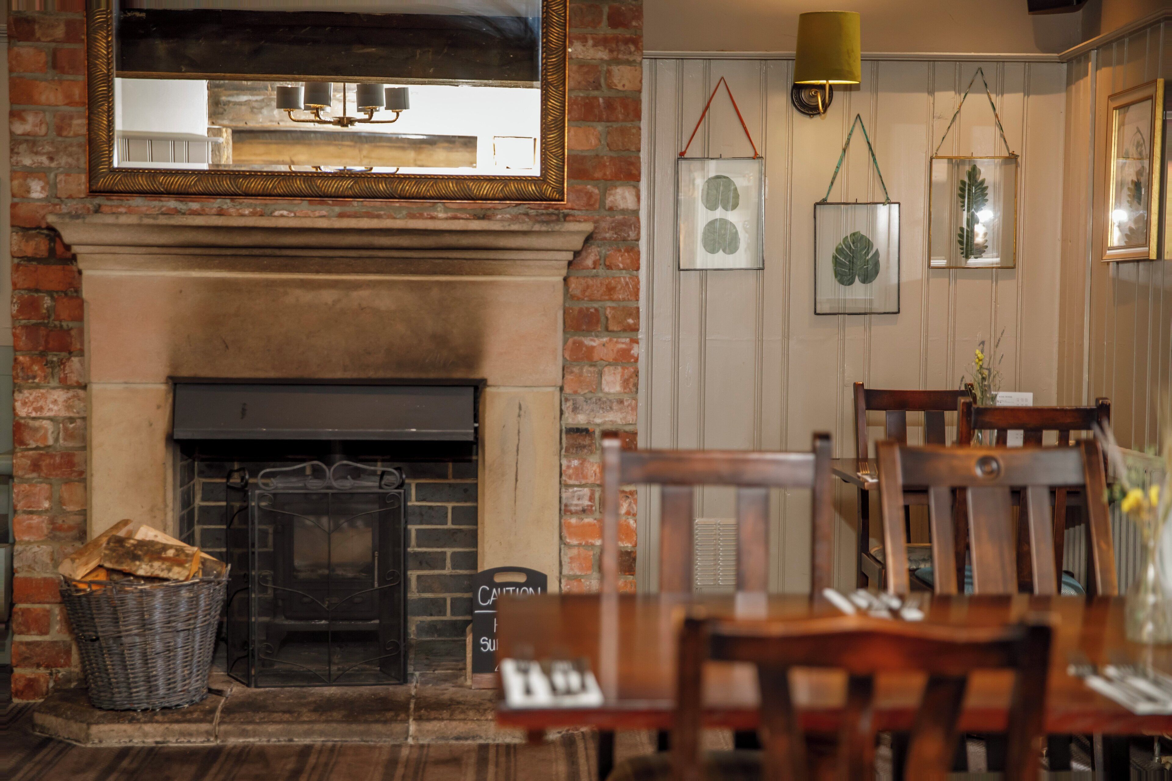 A brick fireplace with a mantel, a dining table with chairs, and framed botanical prints on the wall.
