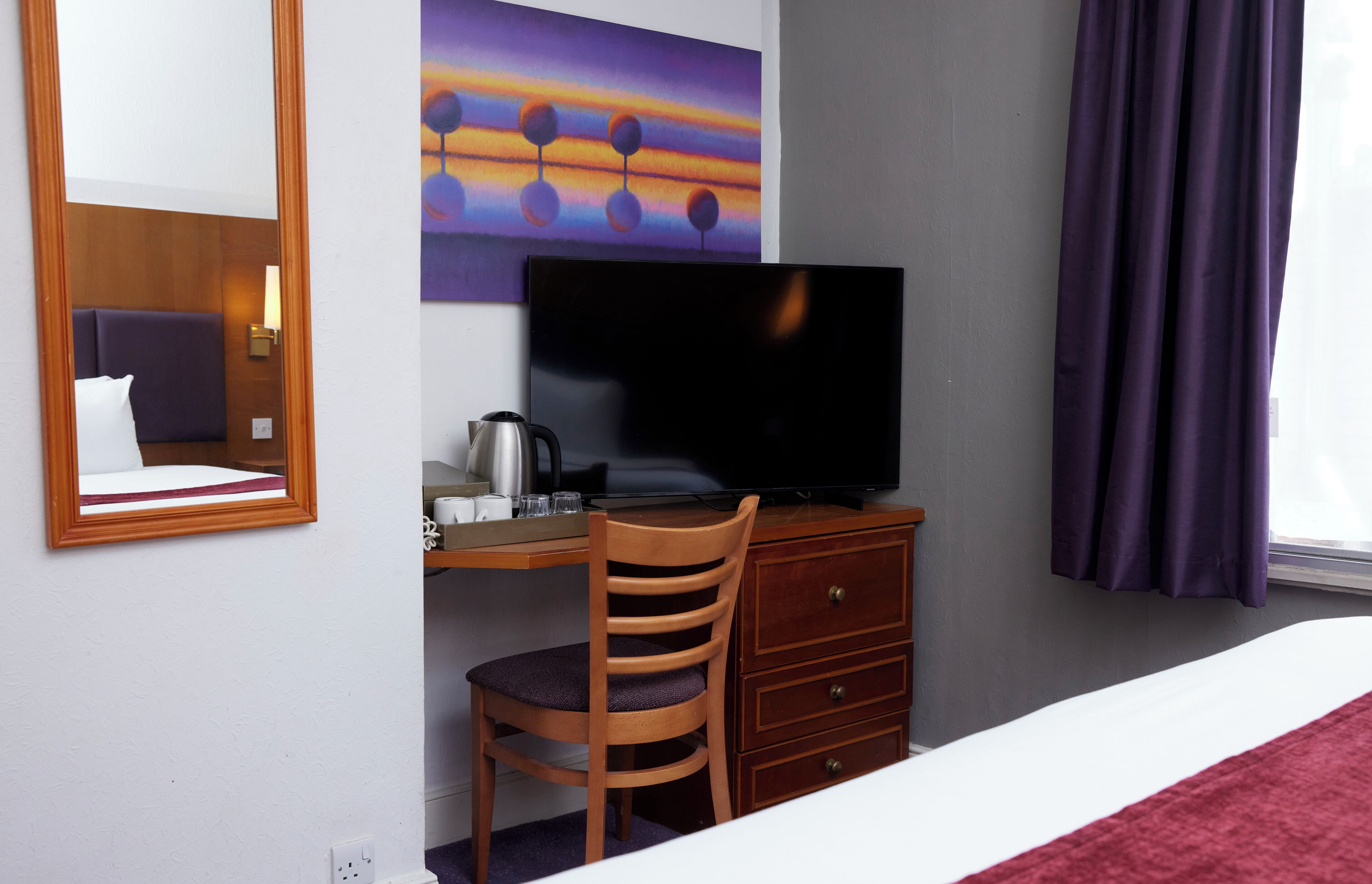 A hotel room with a bed, a desk with a television, a chair, a dresser, and a painting on the wall.