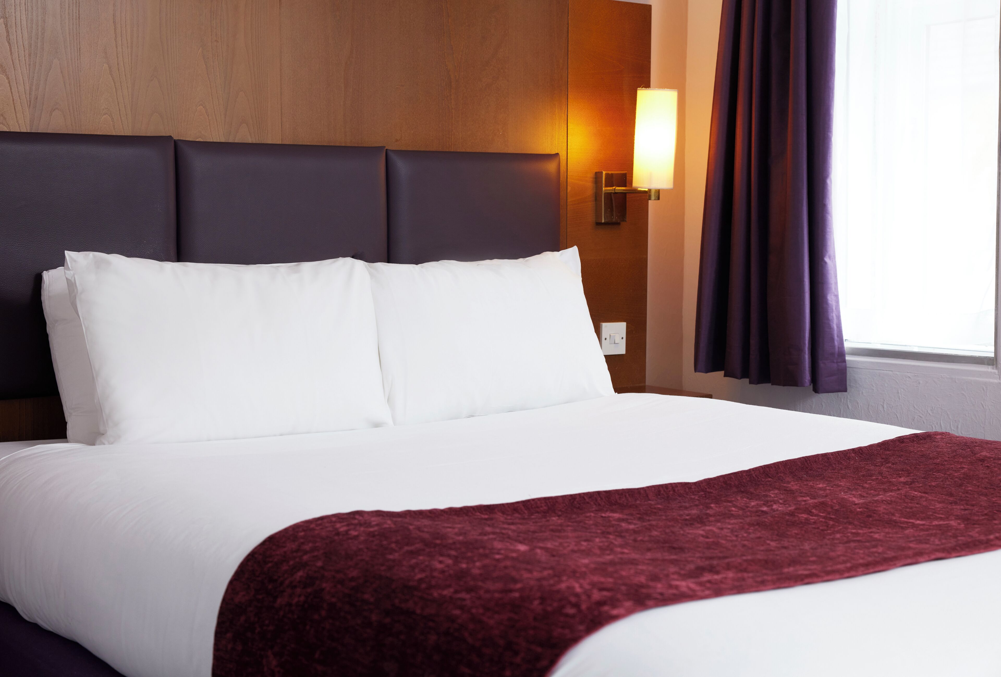 Un lit bien fait, avec du linge de lit blanc et un plaid rouge, une tête de lit sombre et un luminaire fixé au mur.