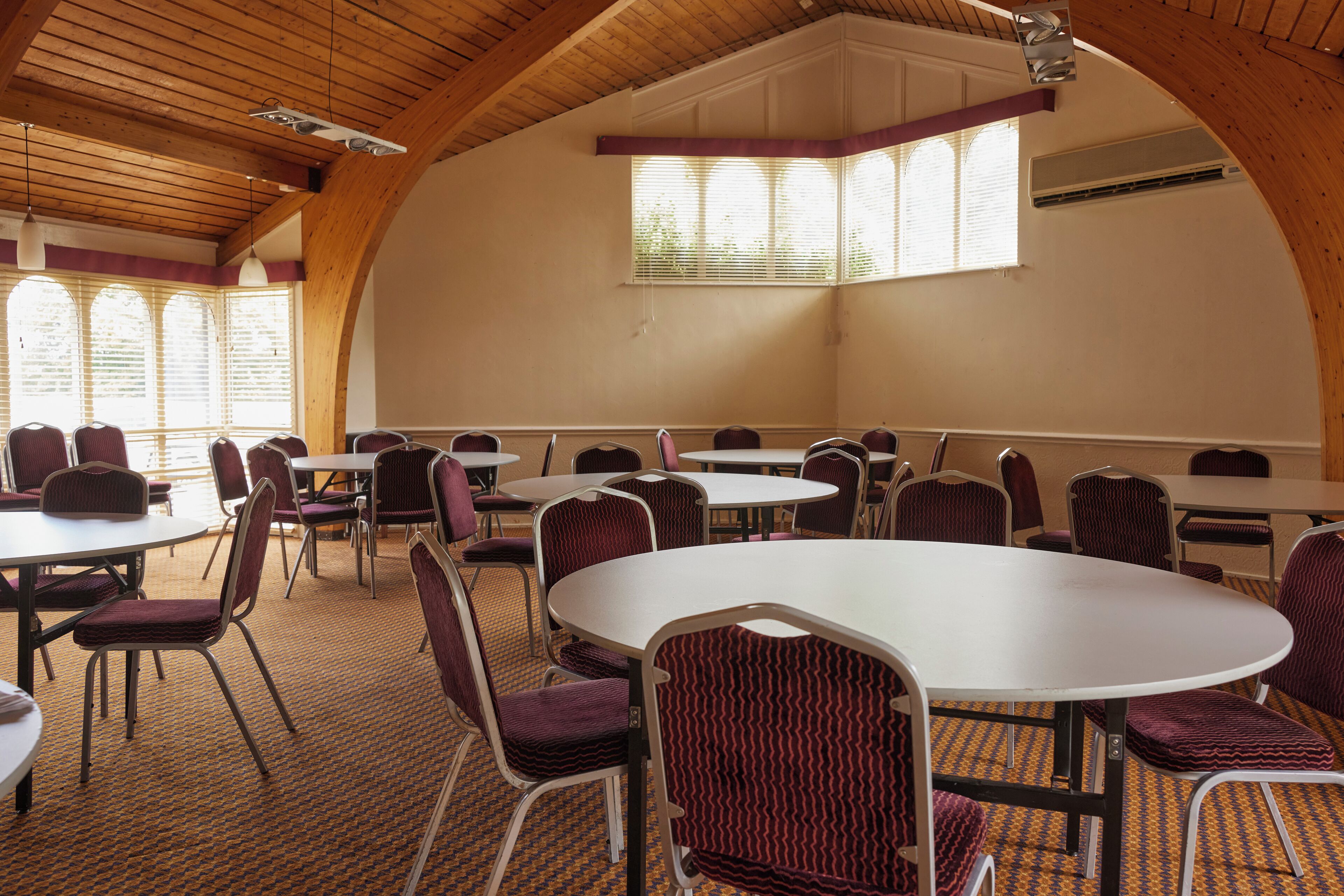 A conference room with round tables and chairs, wooden ceiling beams, and large windows with blinds.