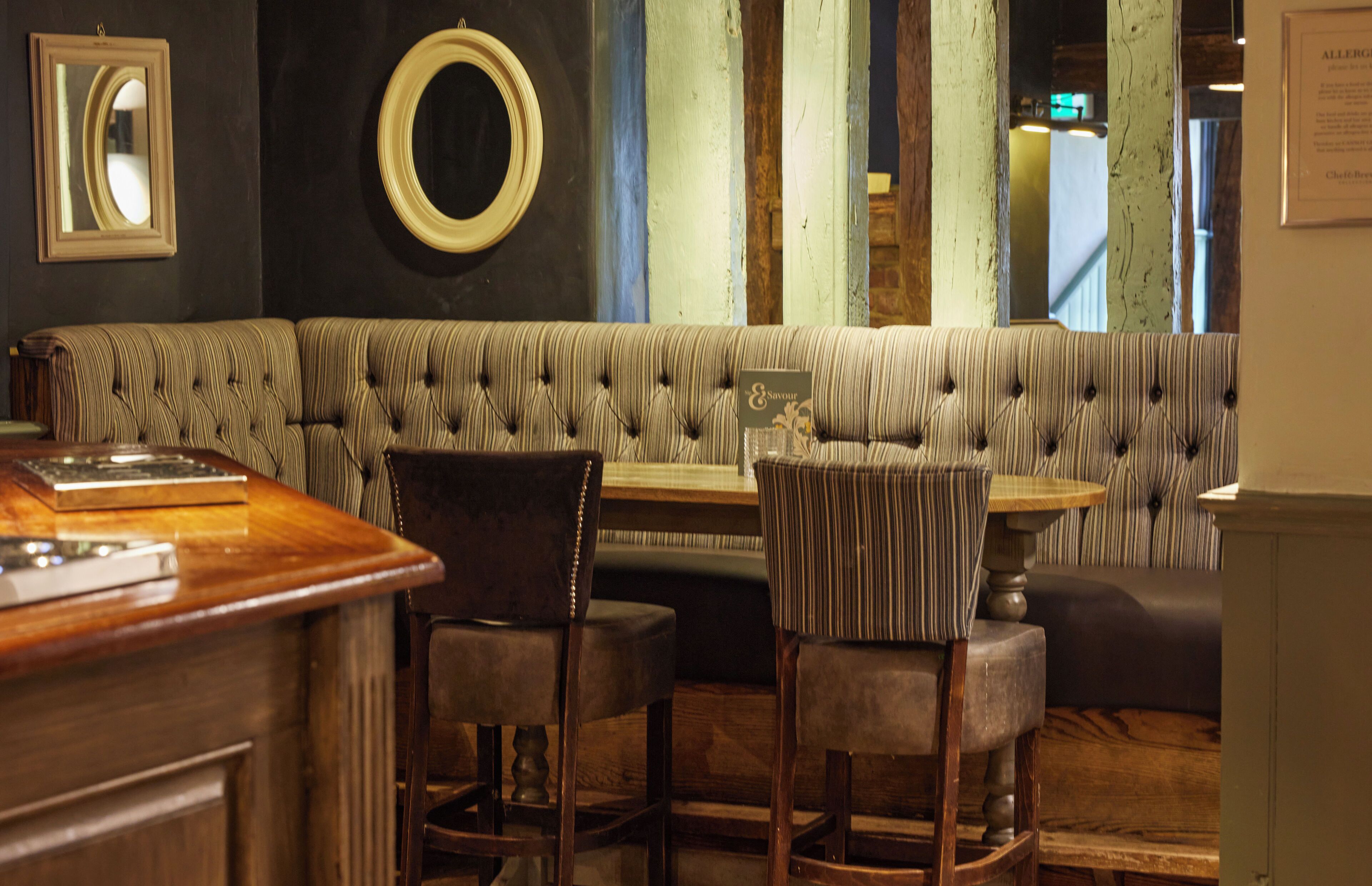 A cozy seating area with a tufted bench and striped upholstered chairs, a wooden bar counter, and framed mirrors on the wall.