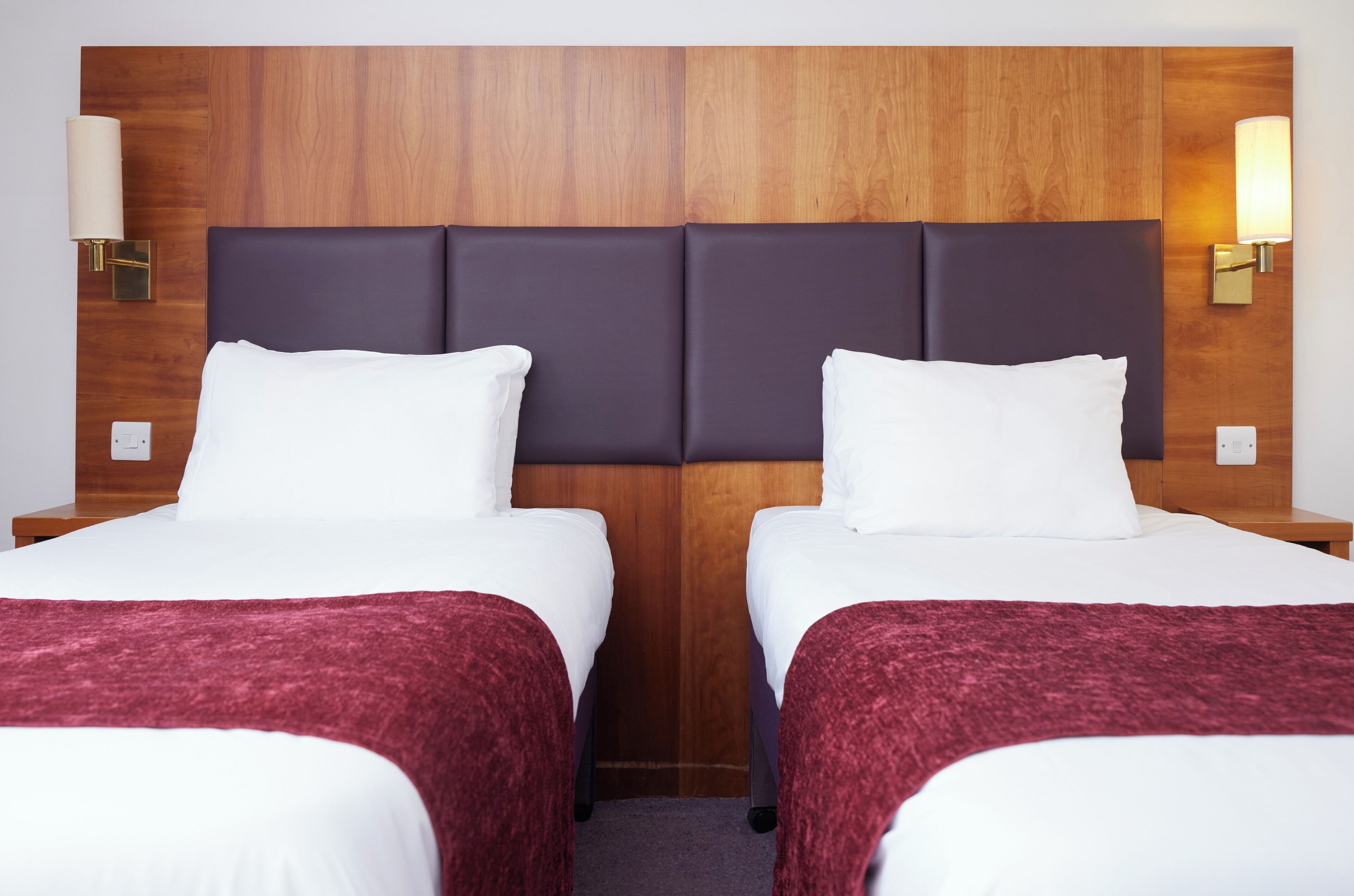 A hotel room with two beds, wooden headboards, and wall-mounted lamps.