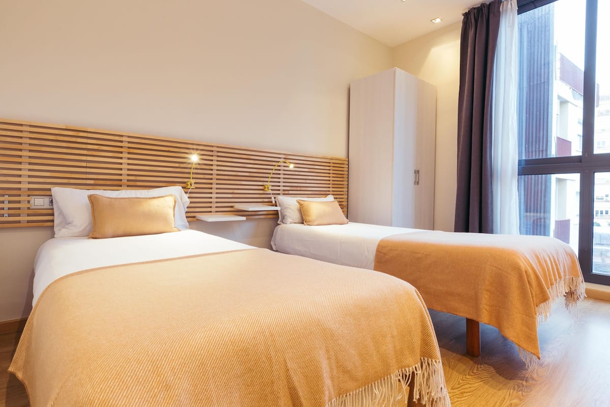 Habitación de hotel con dos camas, cabeceros de madera, un armario y un ventanal con cortinas.