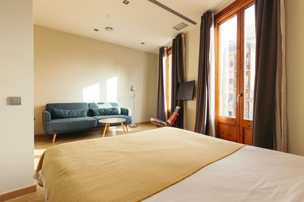 Habitación de hotel con cama, sofá, una mesita redonda, televisor y ventanas grandes con cortinas.