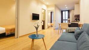 A modern hotel room with a bed, a blue sofa, a small table with a vase, a TV mounted on the wall, and a wooden floor. - Casa Felipa Plaza España (Barcelona)