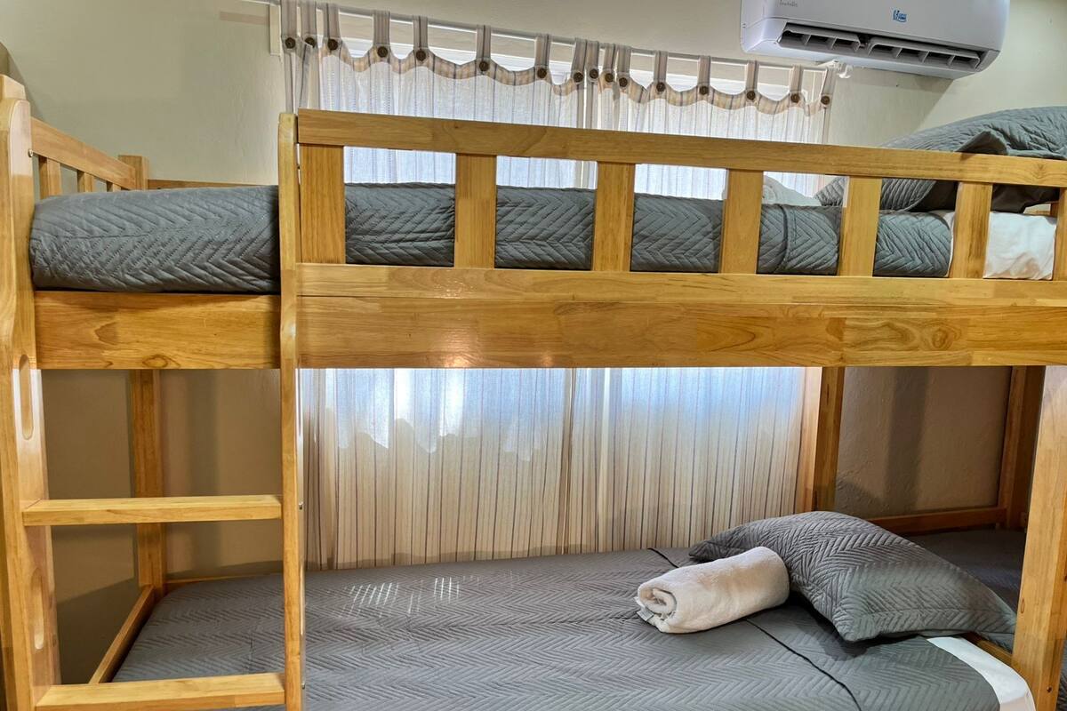 Ein Etagenbett mit Holzgestell, grau gestreuter Bettwäsche und einem Fenster mit durchsichtigen Vorhängen.
