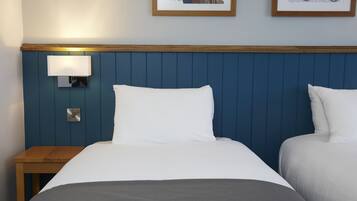 Habitación de hotel con cabecera azul, dos camas, una mesita de noche, una lámpara de pared y cuadros en la pared.