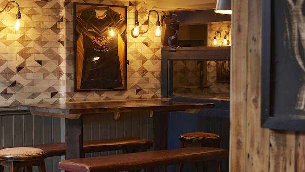 A bar area with wooden stools and benches, a tiled wall with geometric patterns, and pendant lighting.