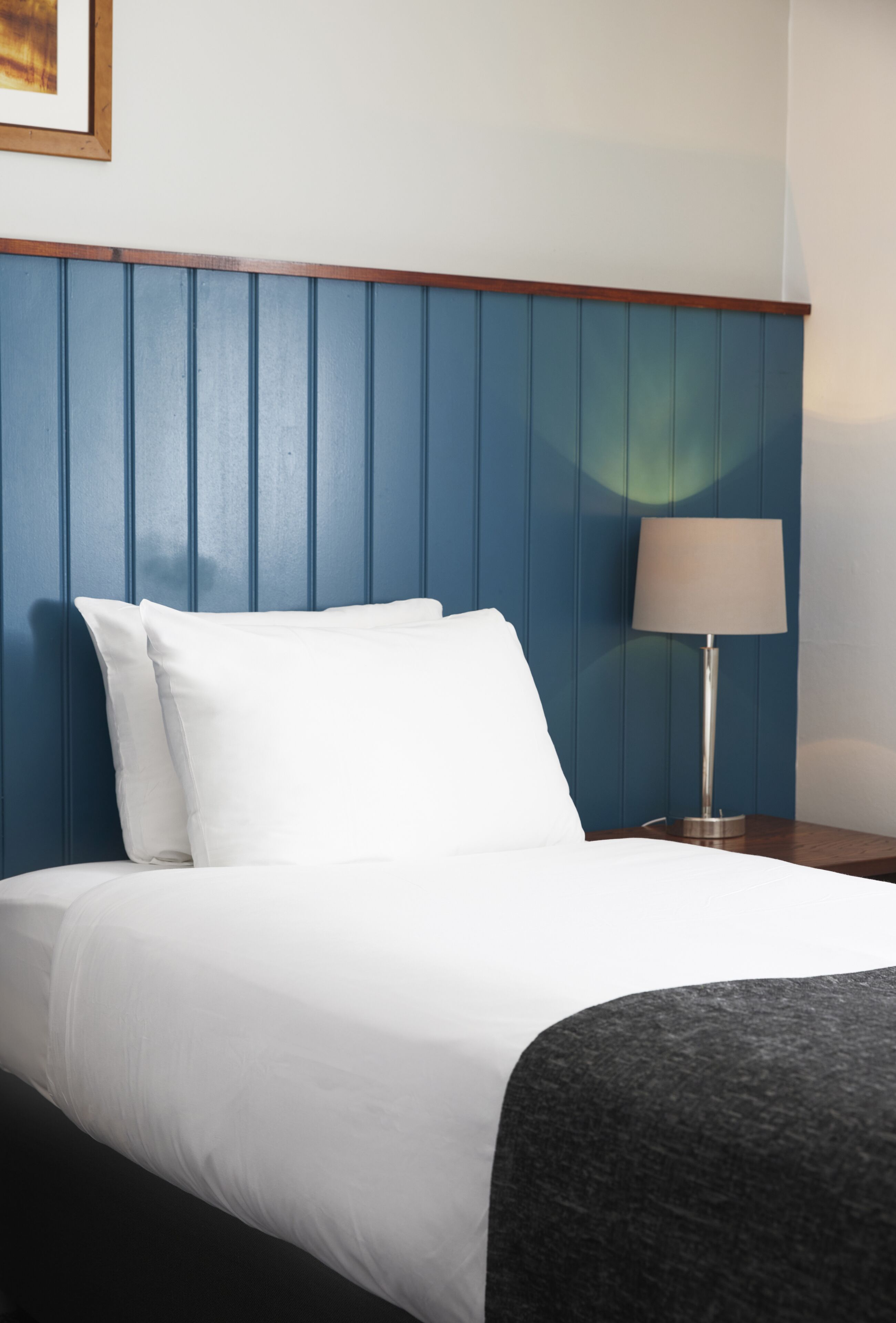 Habitación de hotel con cama, ropa de cama blanca, cabecera azul, lámpara de noche y un cuadro en la pared.
