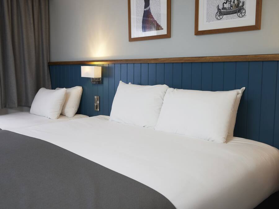 A neatly made bed with white linens and a gray throw blanket. There are two wall-mounted lamps on either side of the bed.