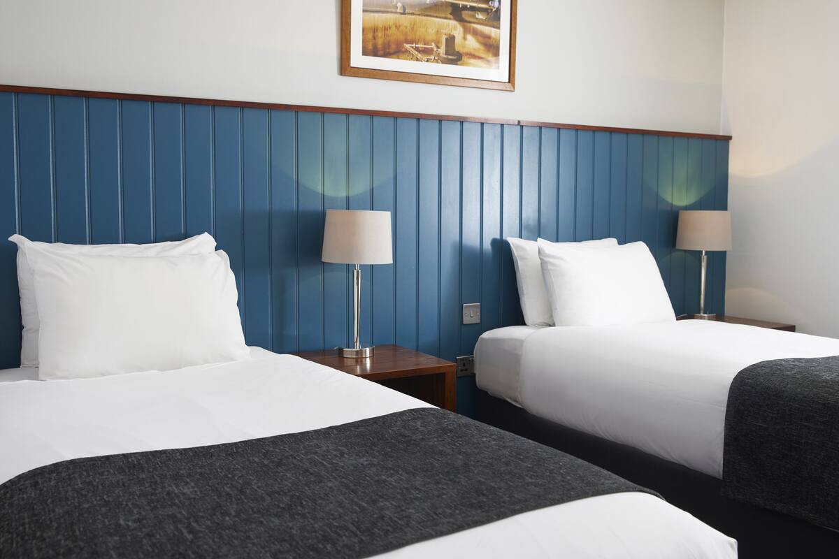 Une chambre d’hôtel avec deux lits, chacun agrémenté de draps blancs et d’un plaid gris. Il y a deux tables de chevet avec des lampes. La chambre est dotée d’une tête de lit bleue à panneaux et d’un tableau encadré au mur.