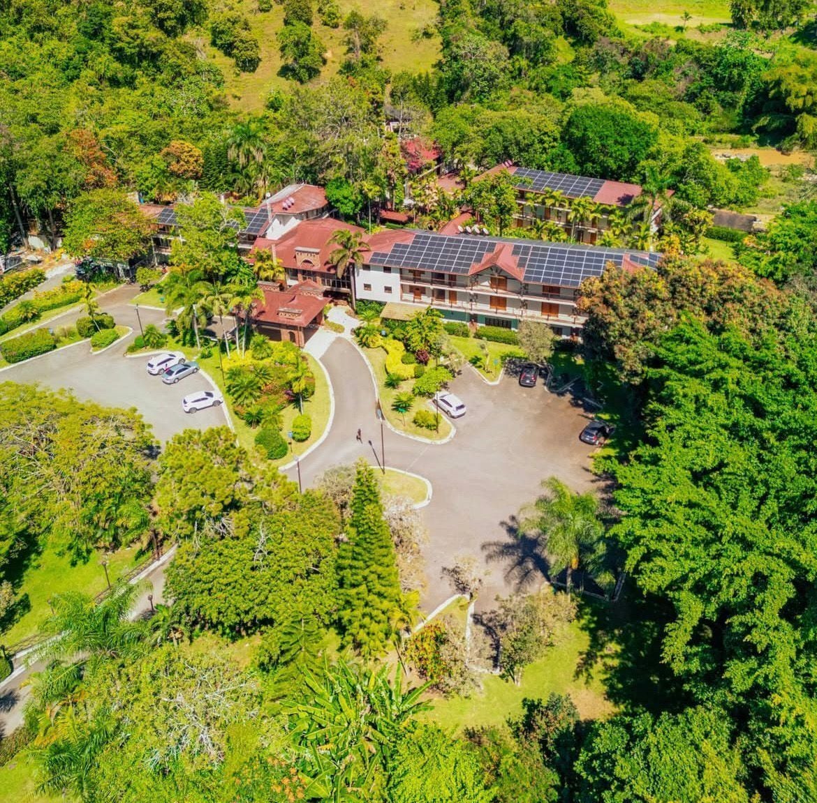 Vista aérea de un hotel con paneles solares en el techo, rodeado de vegetación exuberante y un estacionamiento.