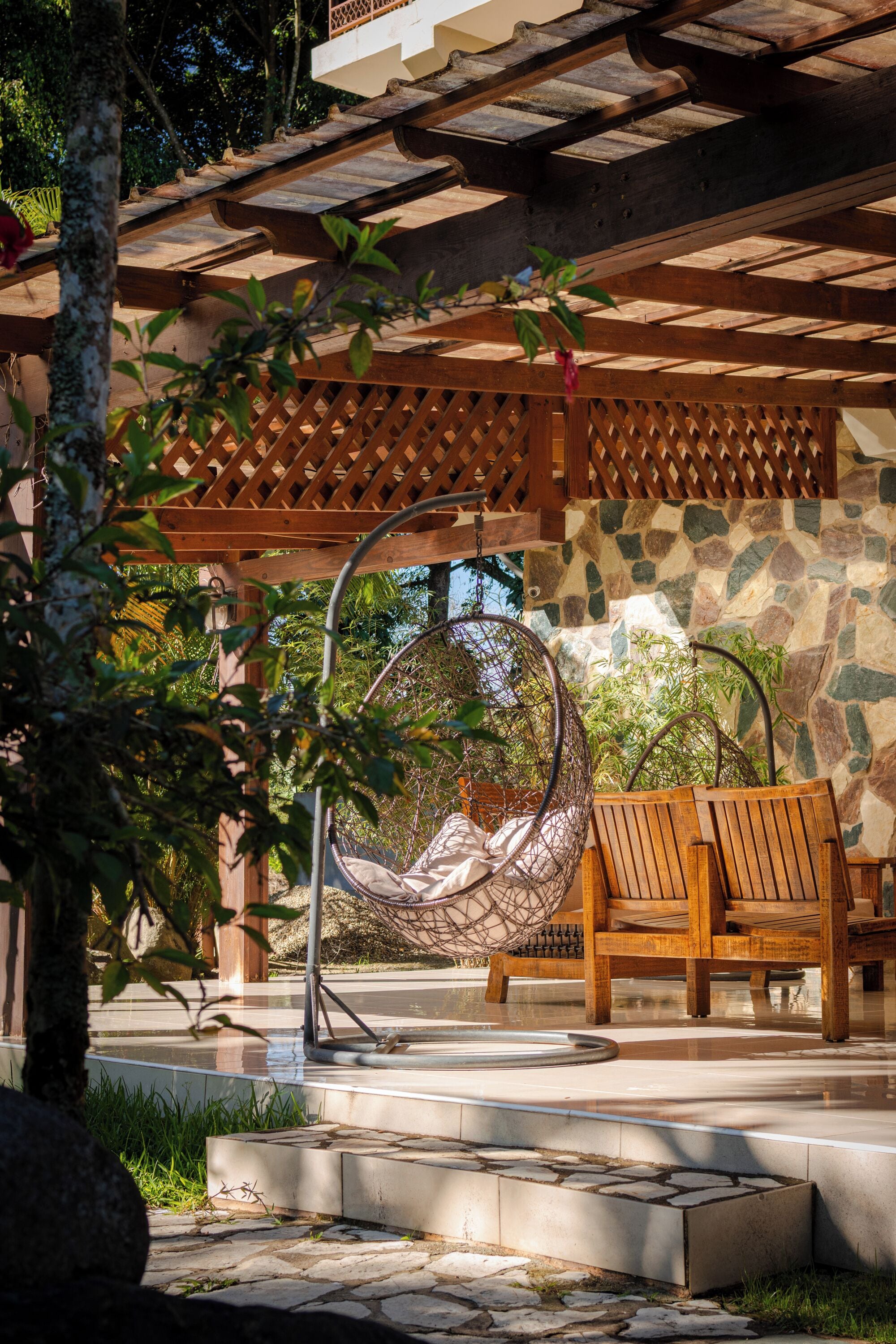 A wooden pergola with a hanging chair and a wooden bench, stone wall, and greenery.