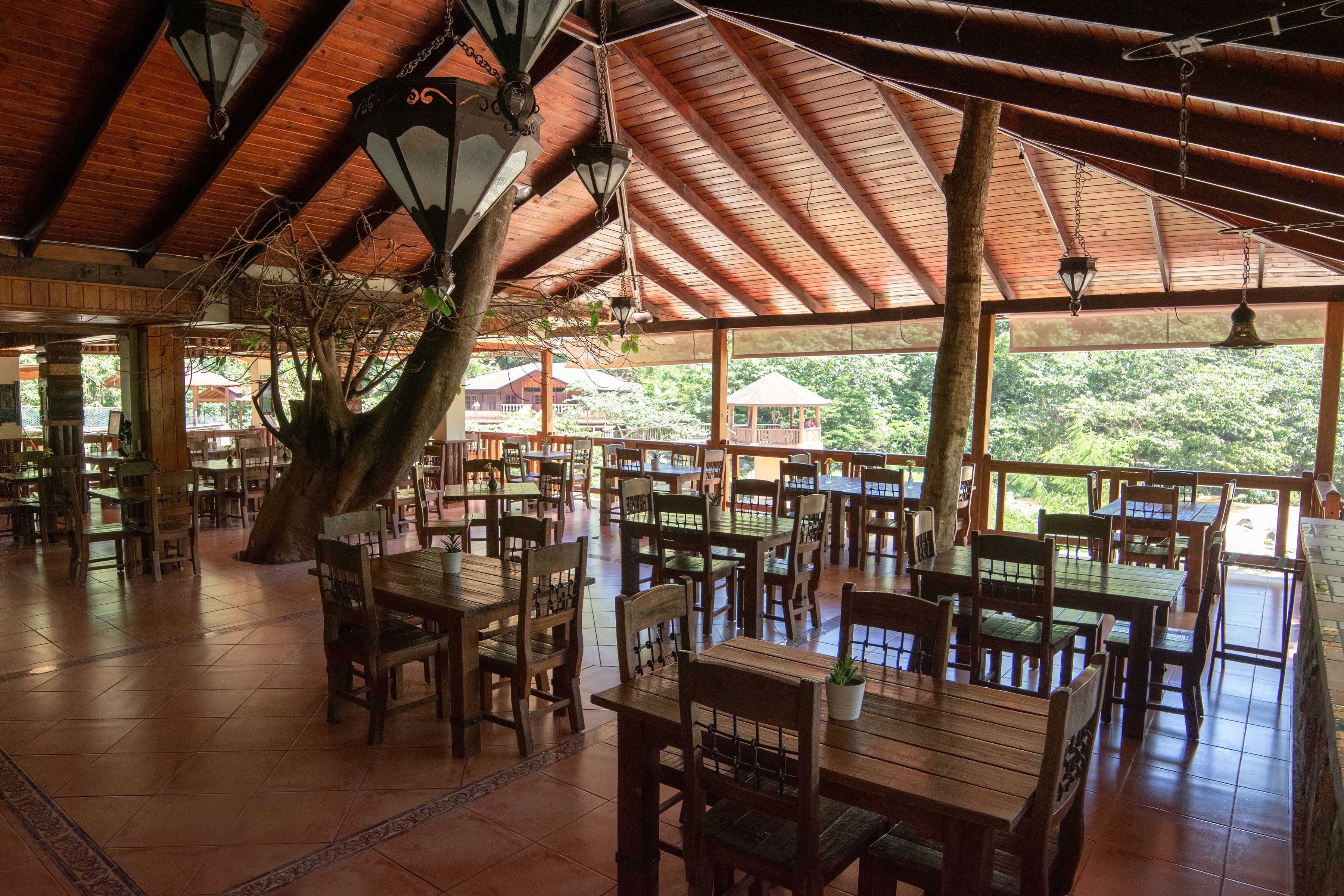 Un comedor amplio con mesas y sillas de madera, ventanas grandes y faroles colgantes.