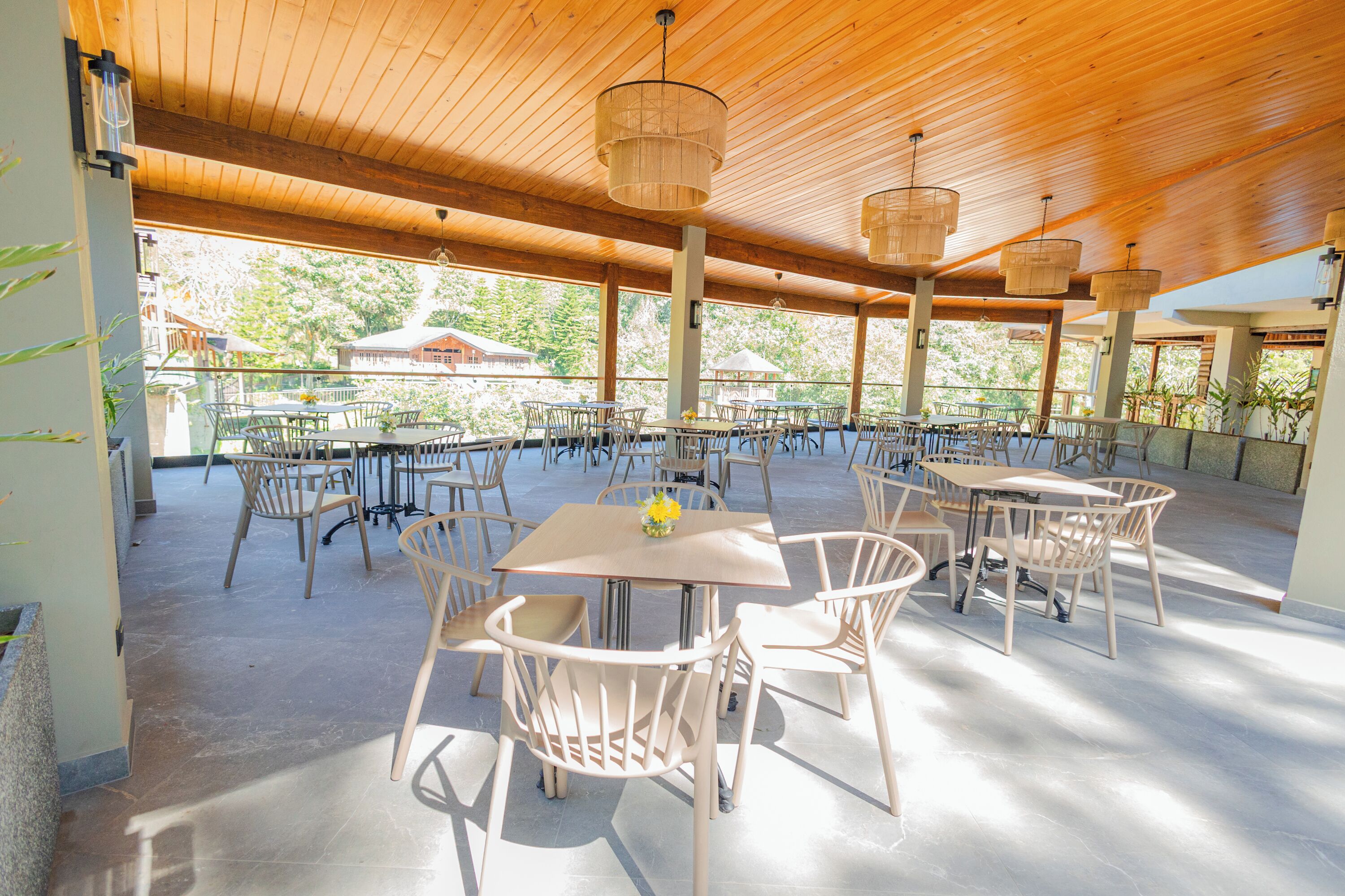 An outdoor patio with multiple tables and chairs, wooden ceiling, and large windows offering views of the surrounding area.