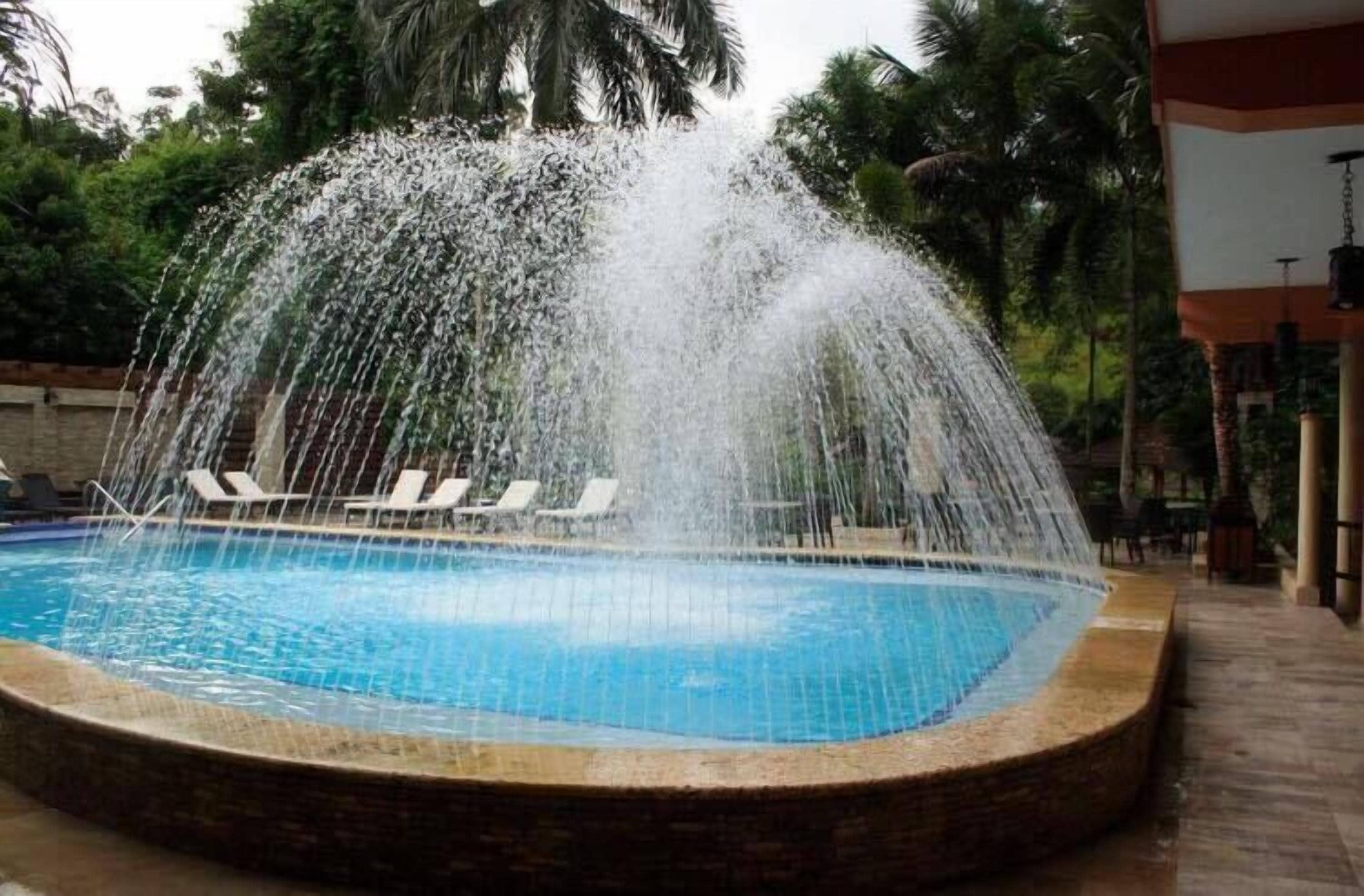 A swimming pool with a water fountain feature, surrounded by lounge chairs and palm trees.