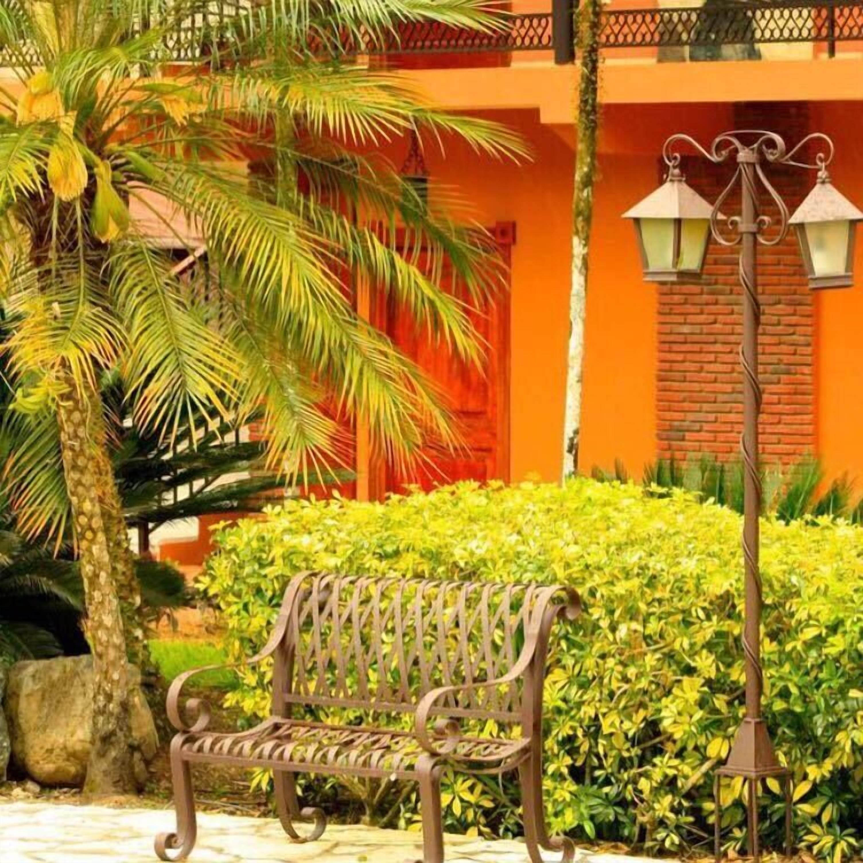 Un banco de hierro forjado, una palmera y un seto en un patio con pared de ladrillo y farola metálica.