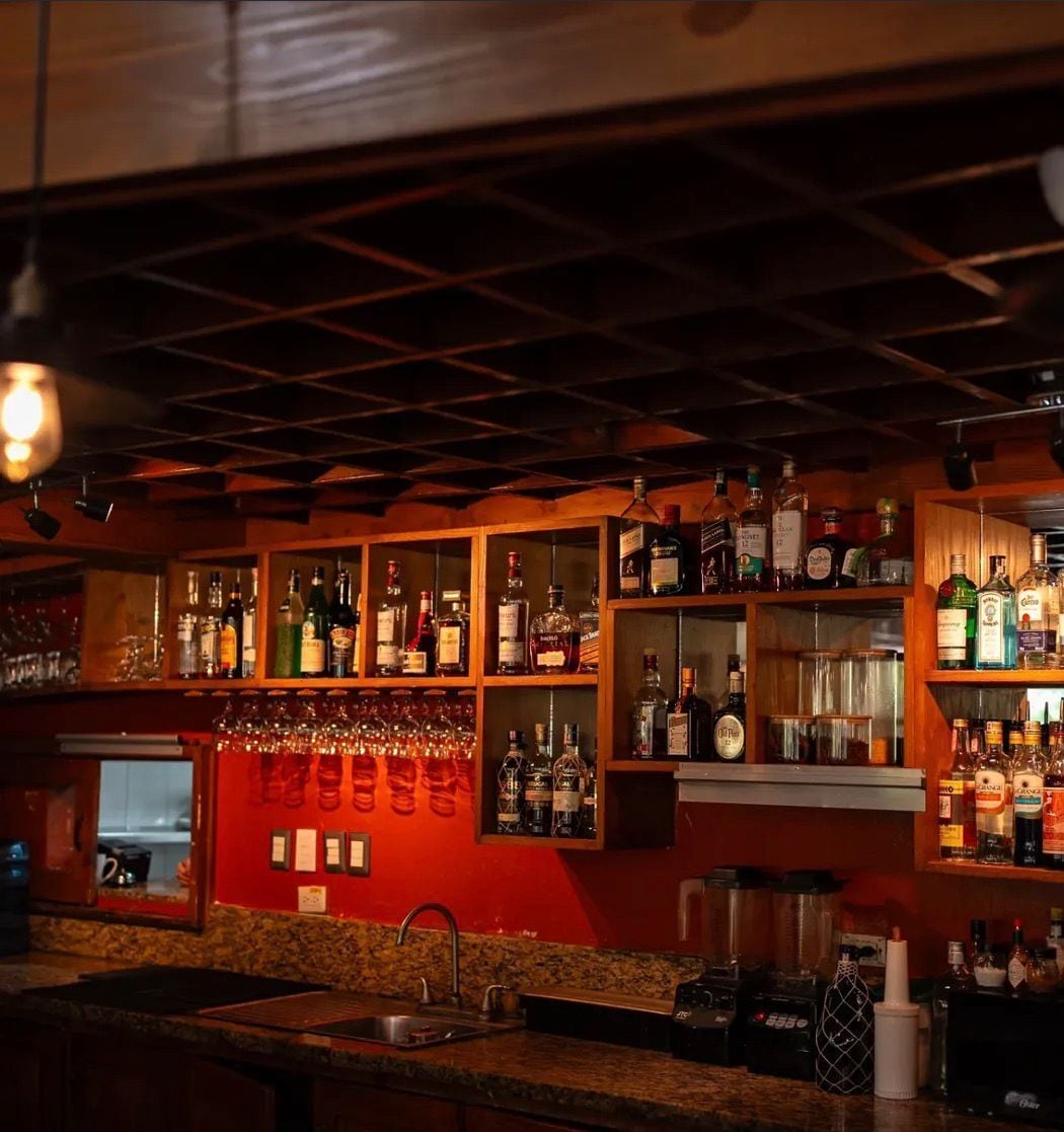 A bar area with shelves of liquor bottles, glasses, and a countertop with a sink and bar tools.