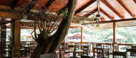 A restaurant with wooden tables and chairs, hanging lanterns, and a large tree trunk in the center.