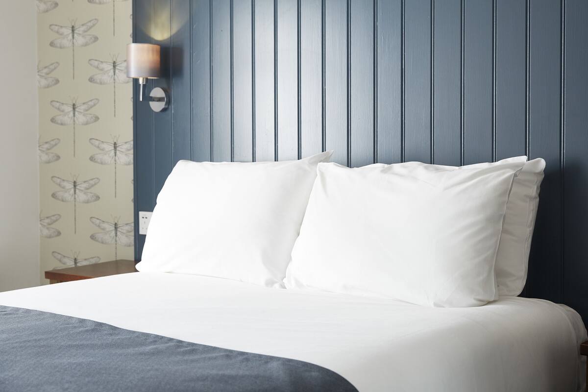 A neatly made bed with white bedding and two pillows, a wall-mounted lamp, and a blue wall with vertical lines.