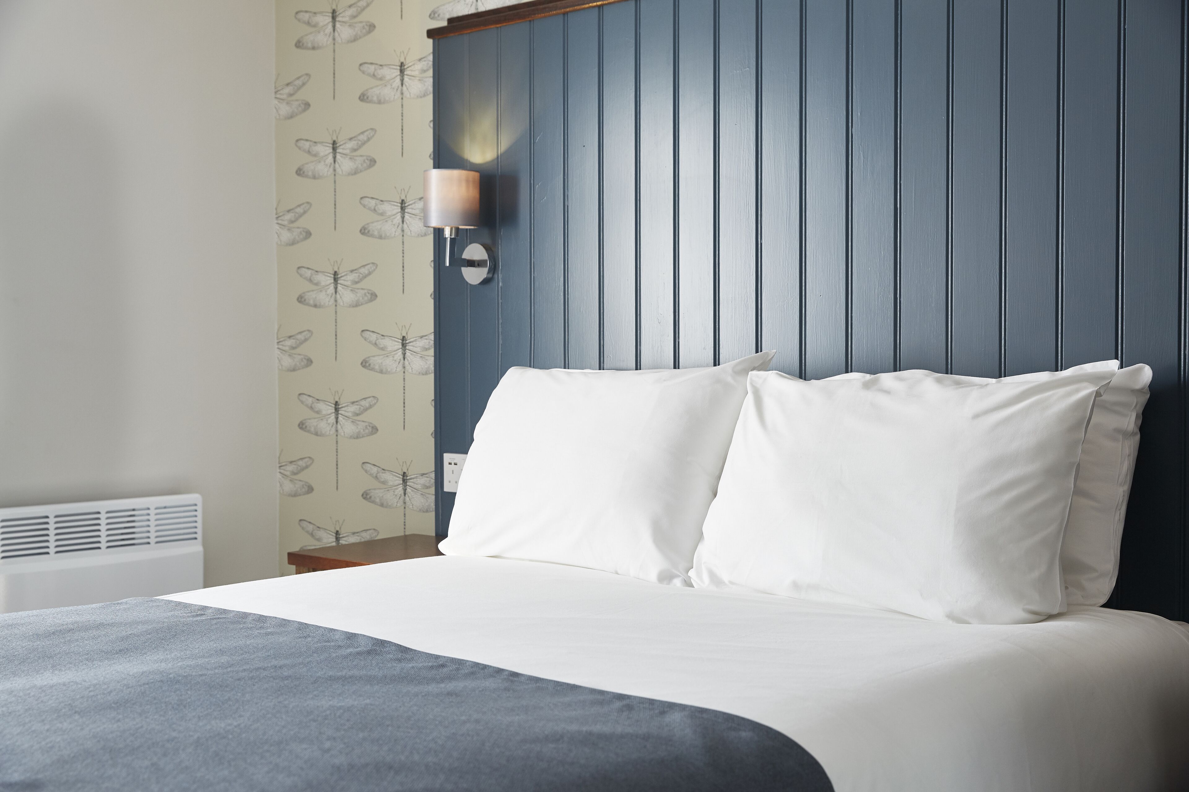 A neatly made bed with white linens in a room with a blue paneled headboard and a wall-mounted light fixture.