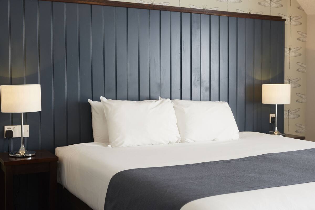 A neatly made bed with white linens and a dark blue headboard. There are two bedside lamps on either side of the bed.