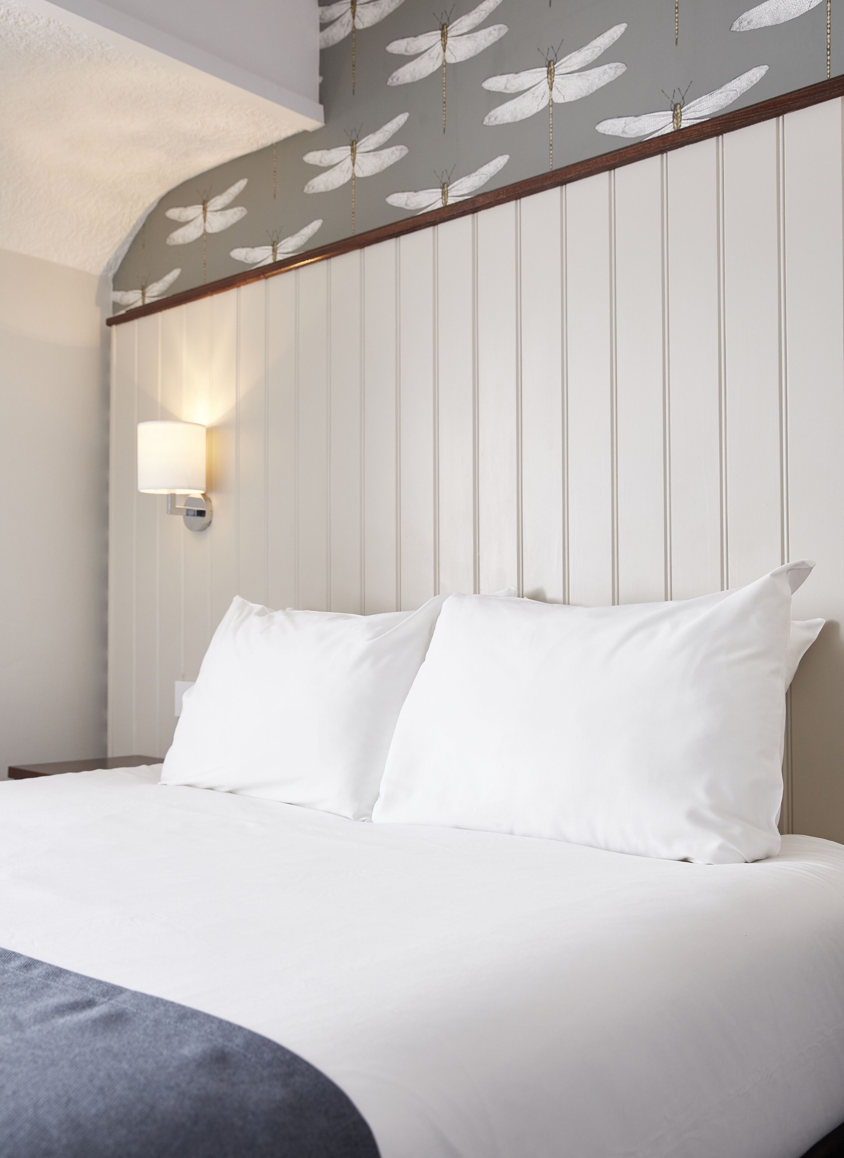 Quarto de hotel com cama, roupa de cama branca, luminária fixada na parede e teto decorativo com padrão de libélula.