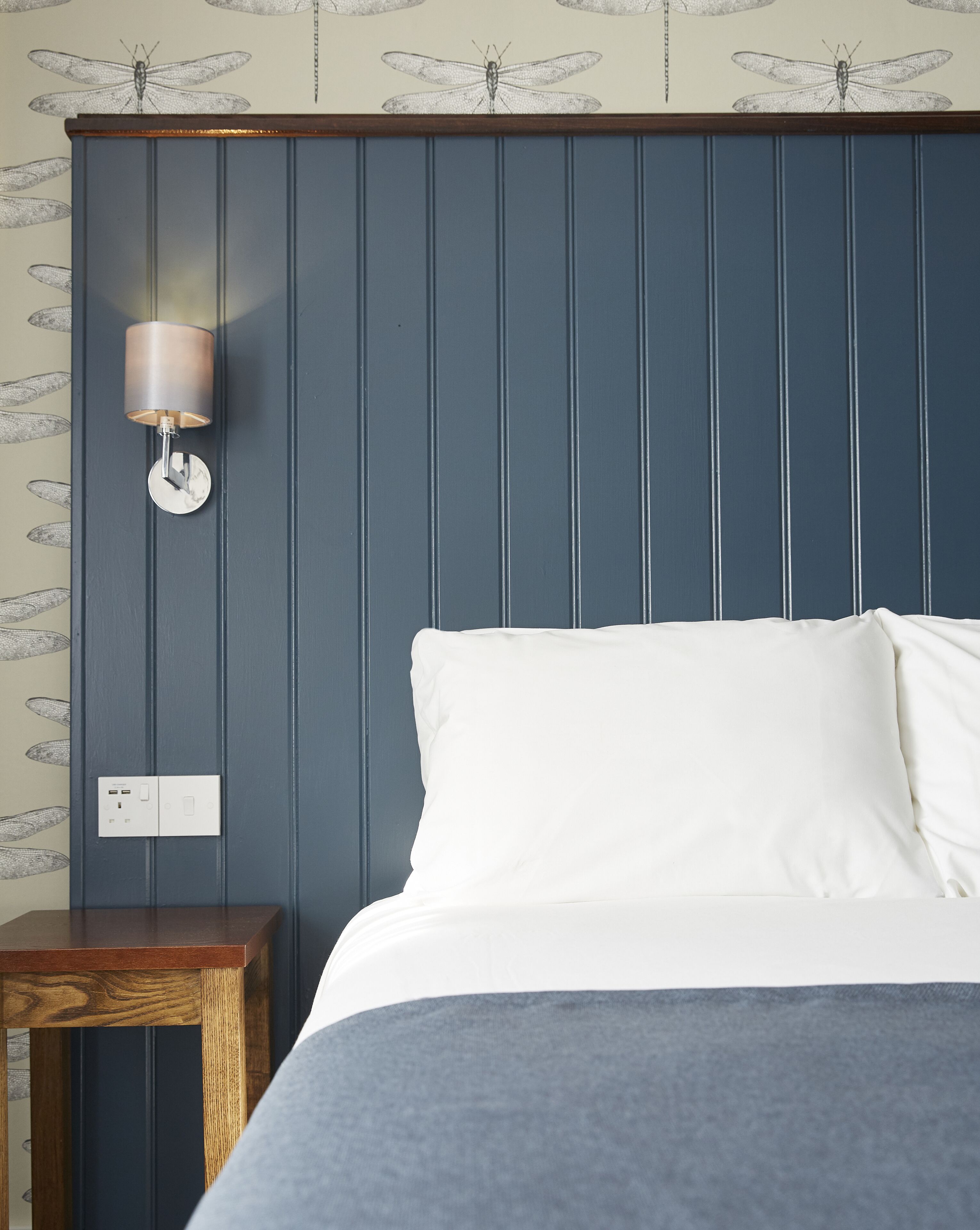 A bedroom with a bed, a nightstand, a wall-mounted lamp, and a power outlet.