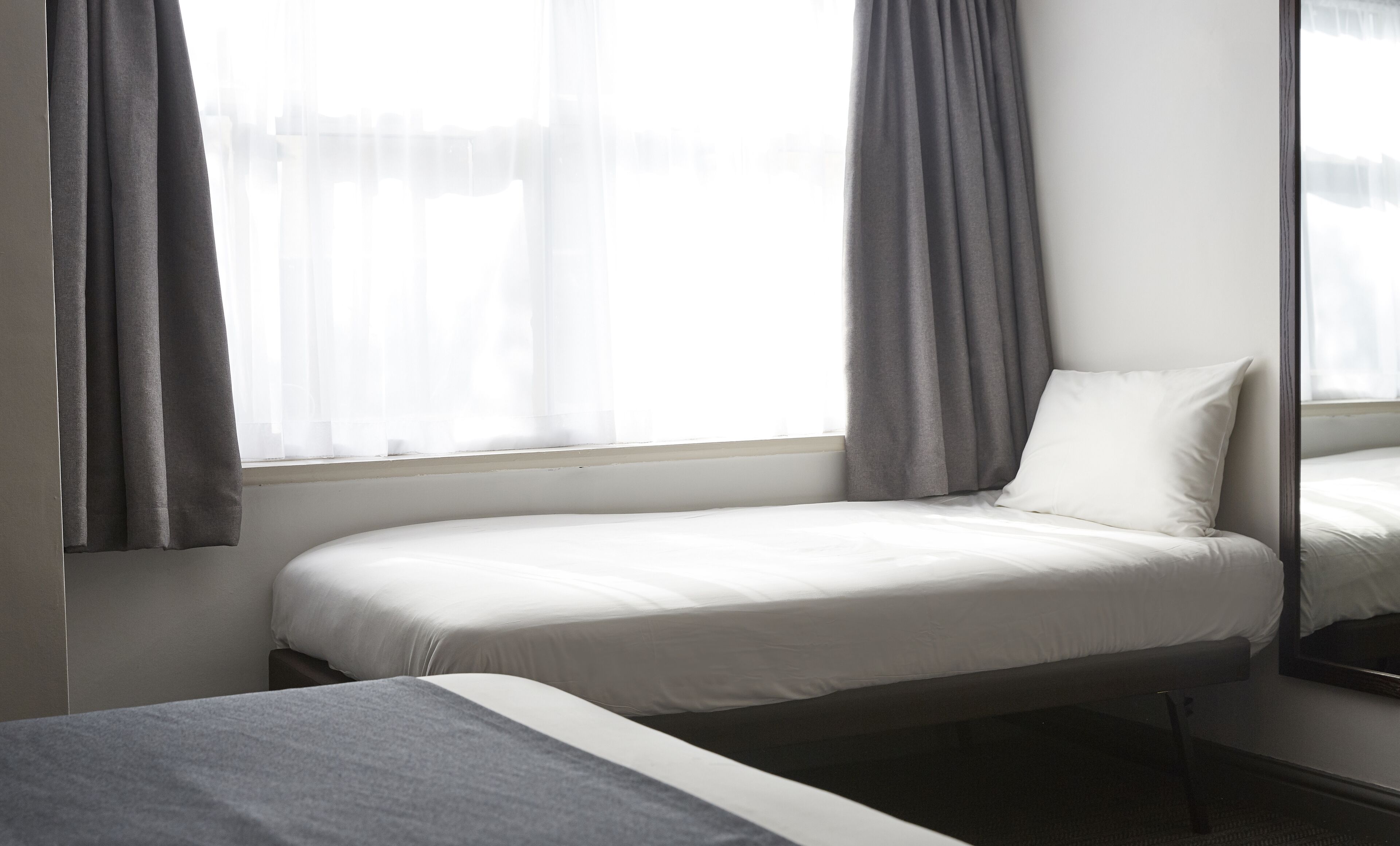A single bed with a white mattress and pillow, grey curtains, and a window with sheer white curtains.