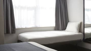 A single bed with a white mattress and pillow, grey curtains, and a window with sheer white curtains.