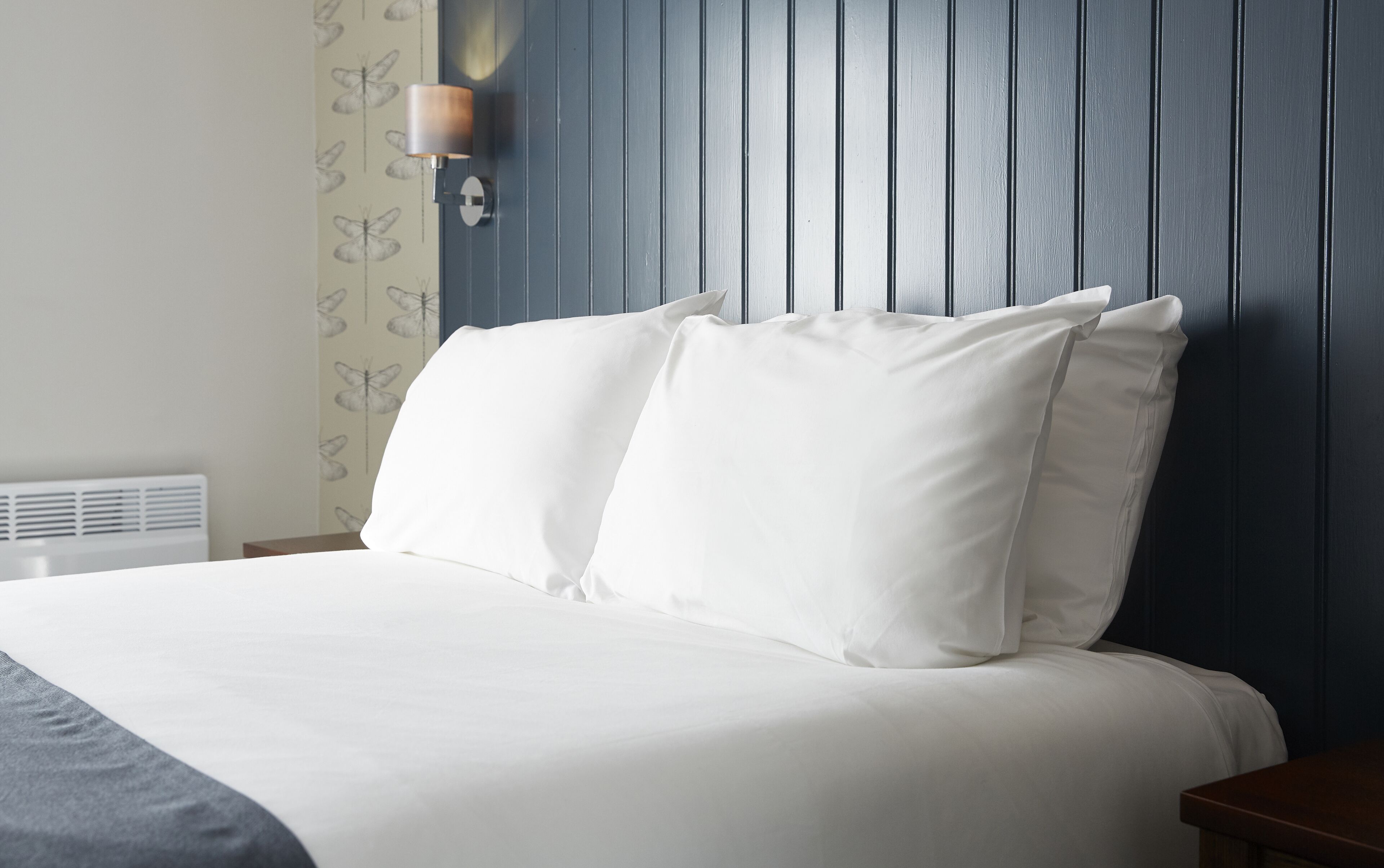 A neatly made bed with white linens, a wall-mounted light fixture, and a blue paneled wall.
