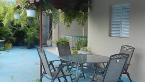 A covered outdoor seating area with a round table and chairs, surrounded by potted plants and a building with a window. - Hotel Navarro (Santo Domingo)