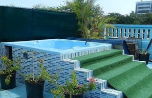 Una piscina en la azotea con revestimiento de azulejos azules, rodeada de plantas en macetas y una escalera que lleva a una zona de césped artificial verde.
