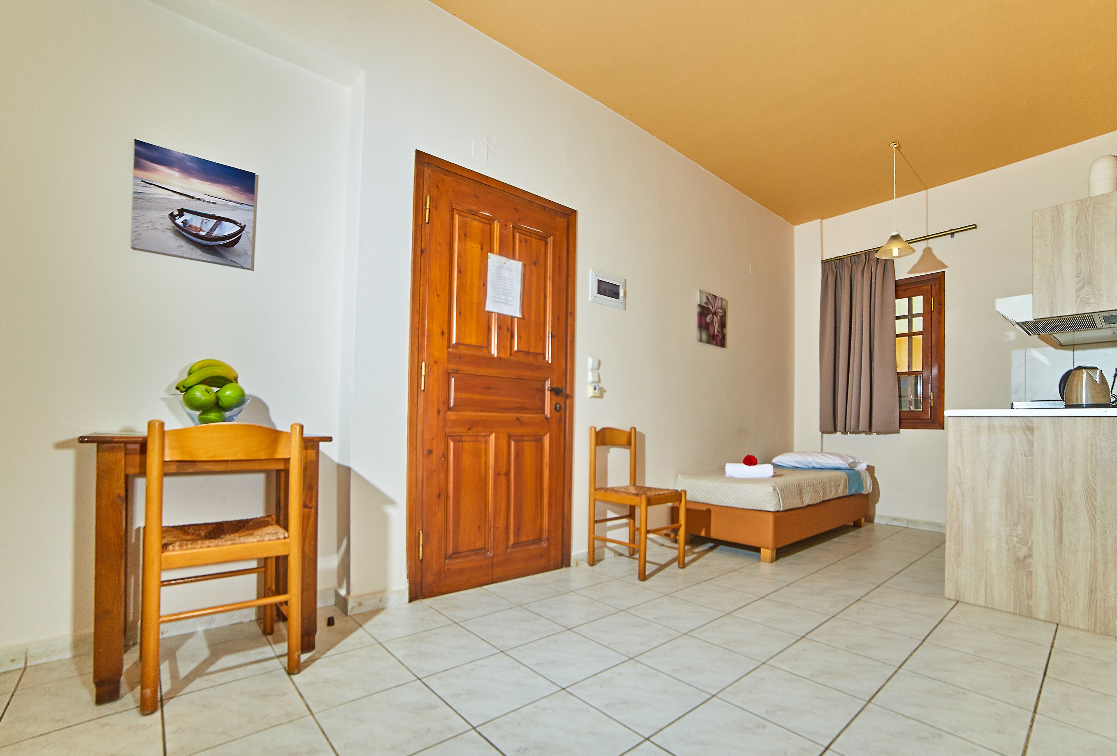 A room with a wooden door, a bed, a chair, a small table with a bowl of fruit, and a picture on the wall.