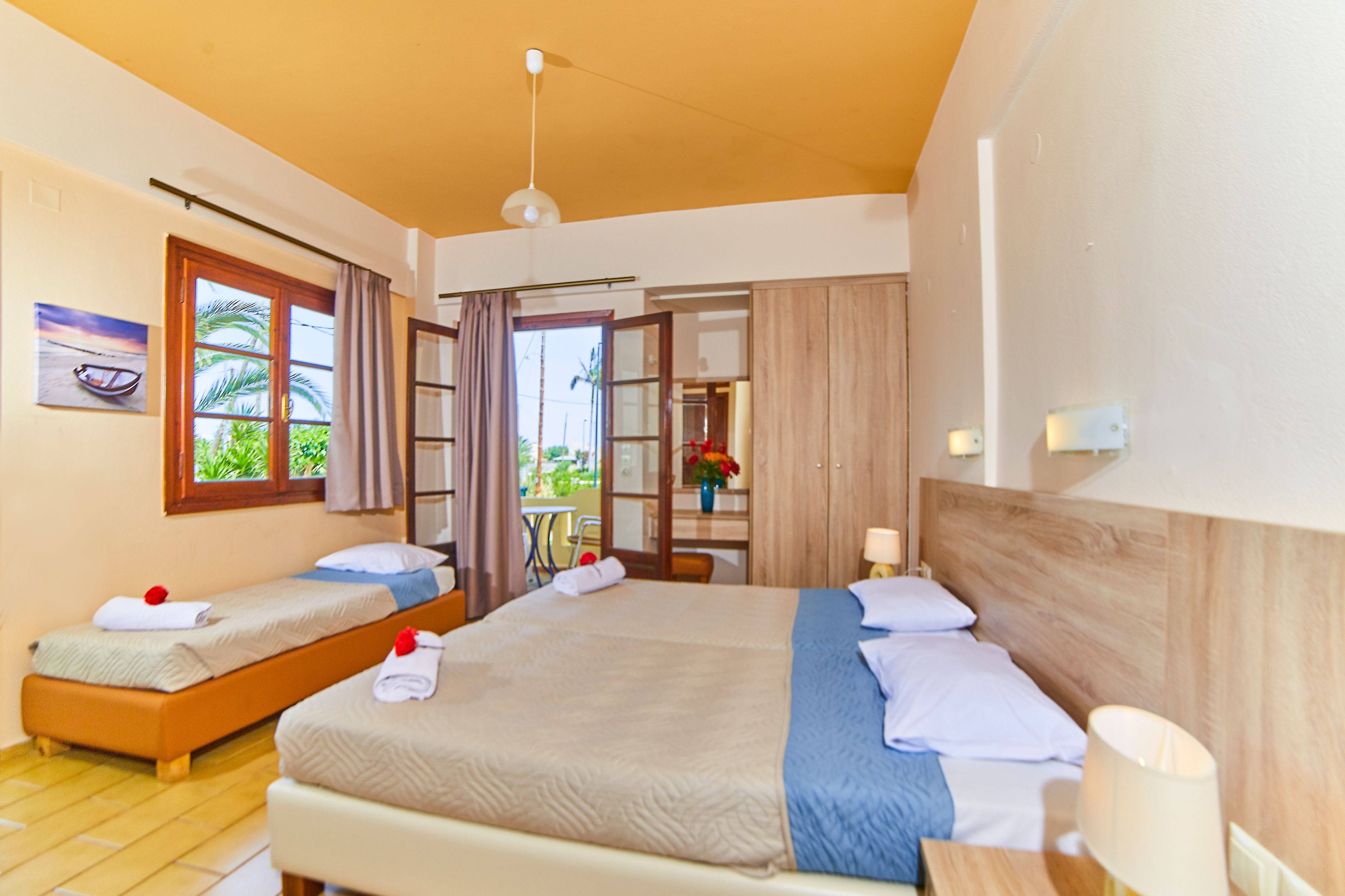 Habitación de hotel con dos camas individuales, una cama grande, cabecero de madera, ventana con vistas y una mesita con lámpara.
